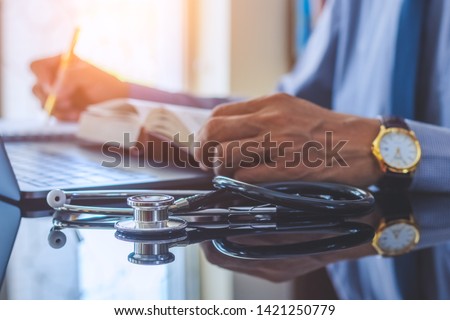 Close up smart male doctor or physician reading text book and writing information,work on laptop computer with medical stethoscope on the desk. Medical technology, online education concept.