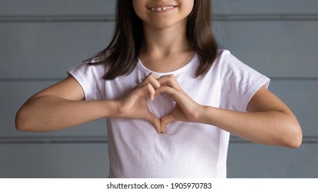 Close Up Smiling Adorable Little Girl Showing Heart Gesture, Happy Cute Preschool Child Kid Wearing White T-shirt Standing On Grey Wooden Wall Studio Background, Charity And Healthcare Concept