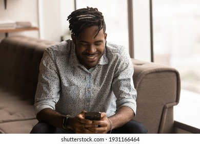 Close up smiling African American man holding phone, browsing apps, sitting on couch, spending leisure time with mobile device, chatting in social network, satisfied customer shopping online