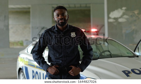 Close up smiling african american young man cops\
stand near patrol car look at camera enforcement happy officer\
police uniform auto safety security communication control policeman\
portrait slow motion