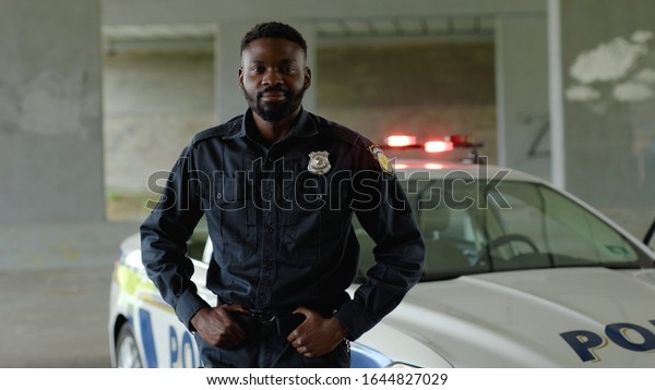 Close up smiling african american young man cops\
stand near patrol car look at camera enforcement happy officer\
police uniform auto safety security communication control policeman\
portrait slow motion