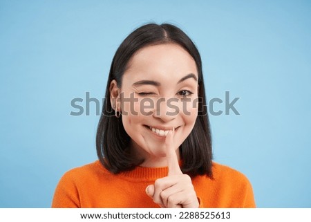 Close up of smiling asian girl, winks at you, tells secret, holds finger on lips, stands over blue studio background.