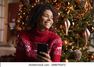 Close up smiling dreamy African American woman holding phone, sitting near Christmas tree at home, looking to aside, enjoying winter holidays with mobile device, shopping online, choosing gifts - Powered by Shutterstock