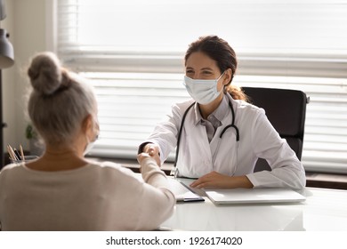 Close up smiling female doctor wearing face mask and uniform with stethoscope shaking mature patient hand at meeting, elderly woman making health insurance deal, elderly generation healthcare - Shutterstock ID 1926174020