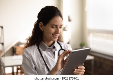 Close up smiling female doctor wearing uniform using tablet, standing in office, browsing medical apps, professional therapist physician working online, chatting, consulting patient, telemedicine - Powered by Shutterstock