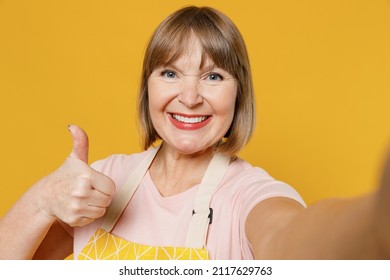 Close Up Smiling Fun Elderly Housekeeper Housewife Woman 50s In Orange Apron Doing Selfie Shot Pov On Mobile Phone Show Thumb Up Isolated Plain On Yellow Background. People Household Lifestyle Concept