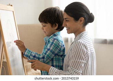 Close up smiling Indian mother with little son drawing on white board with colored chalks, spending leisure time together, having fun, woman teacher baby sitter teaching preschool boy to painting