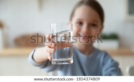 Close up smiling little girl holding glass of pure mineral water, offering to camera, cute pretty child kid recommending healthy lifestyle habit, drinking clean aqua for refreshment