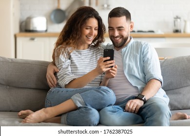 Close up smiling man and woman hugging using smartphone sitting on couch in living room. Loving husband embracing young wife holding smartphone in hand watching funny video at home.