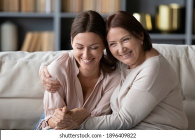 大人の娘を抱き込み、楽しみ、居心地の良いソファに座り、一緒に余暇を楽しむ年配の若い女性と幸せな若い女性、朗報に興奮して接写。