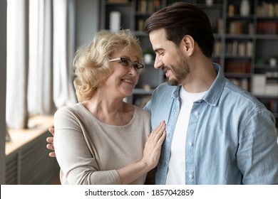 Close Up Smiling Mature Woman And Adult Son Hugging, Looking In Eyes, Family Enjoying Tender Moment, Standing In Modern Living Room At Home, Happy Elderly Mother And Young Man Cuddling