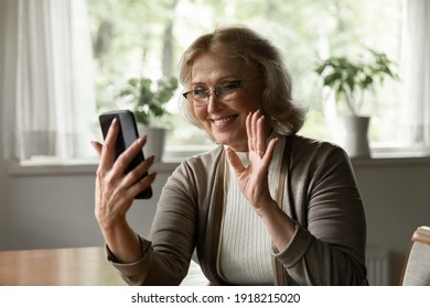 Close up smiling mature woman holding phone, making video call to friends or relatives, chatting online at home, elderly female wearing glasses waving hand, greeting, enjoying pleasant conversation