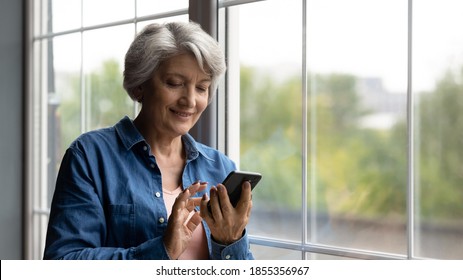 Close up smiling mature woman using smartphone, browsing apps, standing near window at home, senior grey haired female looking at phone screen, chatting or shopping online, enjoying leisure time