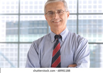 Close Up Of A Smiling Middle Aged Businessman In The Lobby Of A Modern Office Building. Man Is Standing In Front Of A Large Window Wearing A Blue Shirt And Necktie. Horizontal Format.