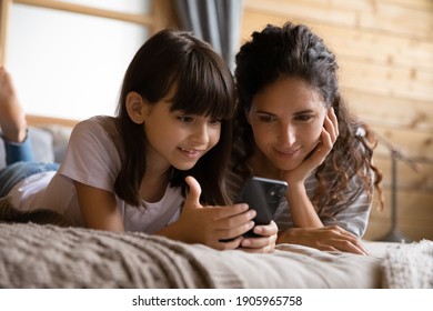 Close Up Smiling Mother And Adorable Daughter Using Phone, Relaxing On Bed At Home, Curious Little Girl Child With Mum Looking At Smartphone Screen, Browsing Apps, Watching Video, Shopping Online