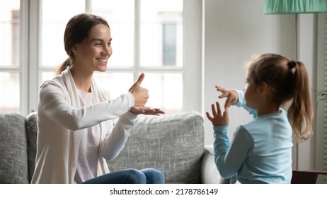 Close up smiling mother and adorable deaf daughter communicating, speaking sign language, sitting on couch at home, therapist teaching little girl, talking nonverbal, hearing disability concept