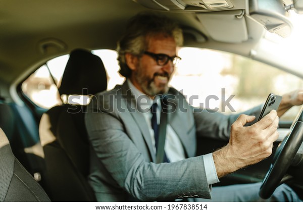 Close up of smiling old\
businessman using smart phone while sitting in his car. Focus on\
phone.