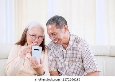 Close Up Of Smiling Old Couple Making A Video Call By Using A Cellphone While Sitting On The Sofa. Shot At Home