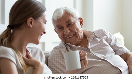 Close up smiling older father and adult daughter chatting, drinking tea or coffee, sitting on cozy couch at home, positive mature grandfather and young woman having fun, talking, sharing news