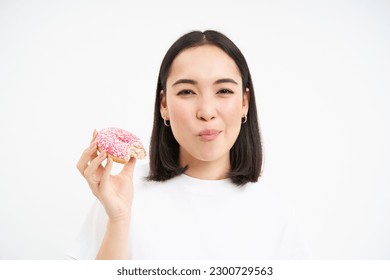 Close up of smiling pleased asian woman, eats glazed pink doughnut, enjoys eating tasty donnut, white studio background.