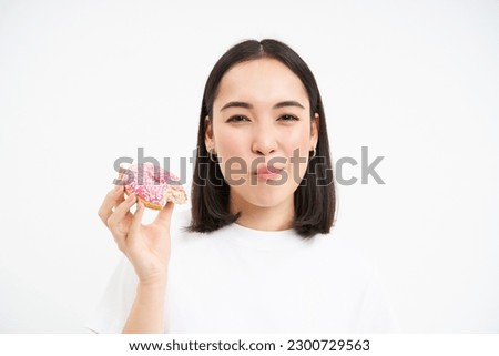 Close up of smiling pleased asian woman, eats glazed pink doughnut, enjoys eating tasty donnut, white studio background.