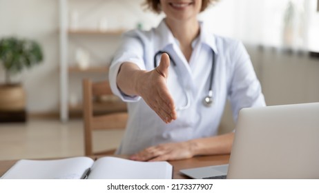 Close Up Smiling Woman Doctor Extending Hand For Handshake To Camera, Physician Therapist Gp Wearing White Uniform With Stethoscope Sitting At Work Desk, Greeting Patient At Medical Appointment