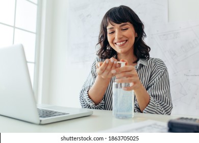 Close up of smiling woman entrepreneur sanitizing her hands sitting at her desk. Cheerful woman following hygiene and covid precautions at workplace. - Powered by Shutterstock