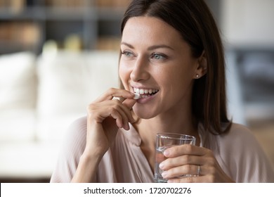 Close up smiling woman taking white round pill, holding water glass in hand, happy young female taking supplement, daily vitamins for hair and skin, natural beauty, healthy lifestyle