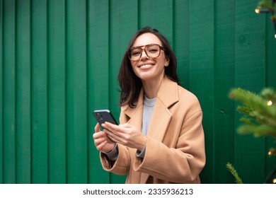 Close up smiling woman using smartphone, standing near Christmas tree, chatting or shopping online, choosing gifts, happy female spending winter holidays with mobile device outside dressed trench coat