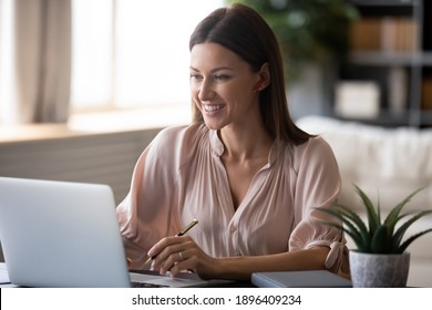 Close up smiling woman wearing wireless earphones using laptop, sitting at work desk at home, happy female watching webinar, studying online, writing notes, businesswoman making video call to partner
