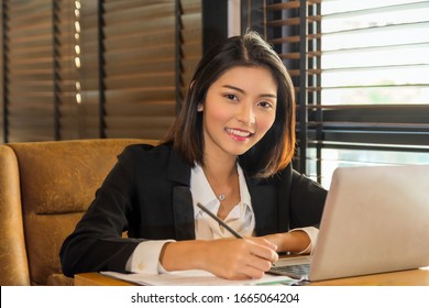Close up smiling young Asian woman in formal suit sitting in front of laptop on wooden table writing note on a book and looking at camera next the winndow at brown tone modern office.
