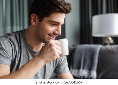 Close up of a smiling young bearded man drinking tea from a mug at the living room - Powered by Shutterstock