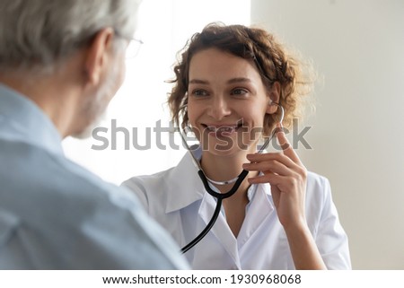 Close up of smiling young Caucasian female doctor use stethoscope examine mature patient heart rate in clinic. Happy caring woman nurse hold phonendoscope listen to elderly man heartbeat in hospital.