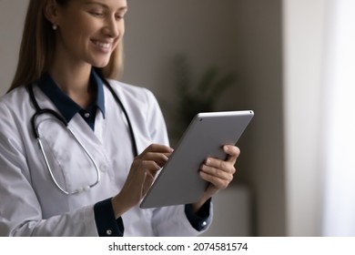 Close Up Smiling Young Female Doctor In Uniform Using Tablet, Standing In Hospital Office, Browsing Medical Apps, Positive Woman Nurse Practitioner Consulting Patient Online, Looking At Device Screen