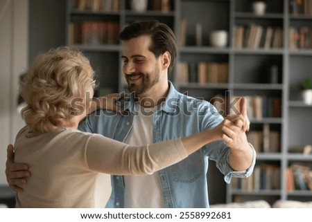 Close up smiling young man dancing waltz with mature mother, standing in modern living room at home, middle aged woman and adult grown-up son enjoying leisure time, family bonding, two generations