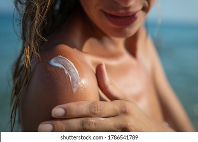 Close up of smiling young woman is applying a sunscreen or sun tanning lotion on a shoulder to take care of her skin on a seaside beach during holidays vacation.