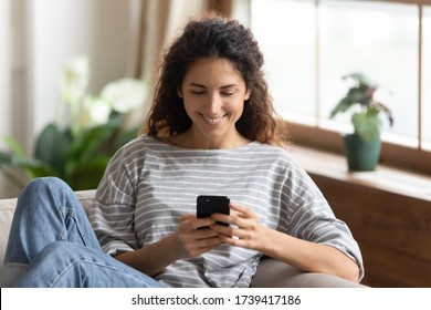 Close up smiling young woman looking at smartphone screen, chatting in social network or surfing internet, shopping online, having fun with phone, sitting on cozy sofa in living room at home