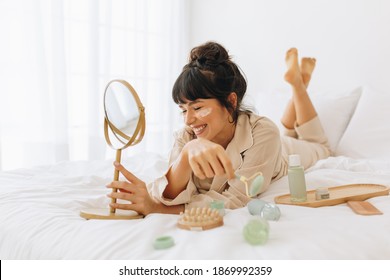 Close Up Of Smiling Young Woman Lying On Bed And Applying Face Cream. Woman Doing Beauty Care Face Massage At Home.