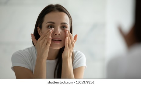 Close up smiling young woman wearing white t-shirt doing facial massage, applying moisturizing cream on under eye skin, looking in mirror, standing in bathroom, enjoying skincare procedure