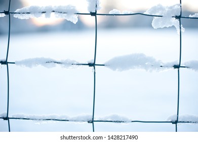 Close Up Of Snow Laying On The Squares Of A Wire Farm Fence With Snow In Background And A Faint Orange Rising Sun - Image