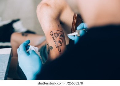 Close Up Soft Focus Shot Of Professional Tattoo Artist Tattooing Funny And Hilarious Pizza Art On Arm Of Young Hipster Man, Wears Hygienic Blue Latex Gloves