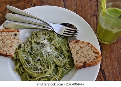 Close up of spaghetti pasta in pesto sauce with toasted bread and mint lemonade, placed on a wooden table and accompanied by stainless steel cutlery