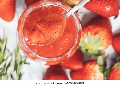 Close up of a spoon is scooping up a fresh strawberry jam from a glass jar. There are several strawberries on the table - Powered by Shutterstock