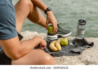 Close up sport man is sitting on cliff near sea and eating green apple after morning cardio.