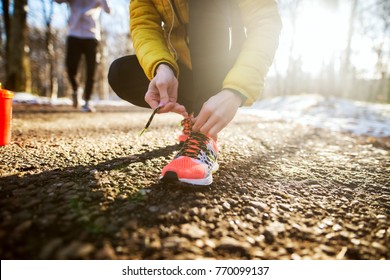 Close Up Of Sporty Active Slim Woman In Sportswear Kneeling On The Road And Tying Shoelaces In The Sunny Winter Morning Outside In Nature With A Trainer Behind Her.