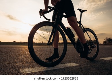 Close Up Of Sporty Young Man With Strong Legs Standing On Road With Black Bike. Natural Landscape, Evening Time. Cycling Concept.