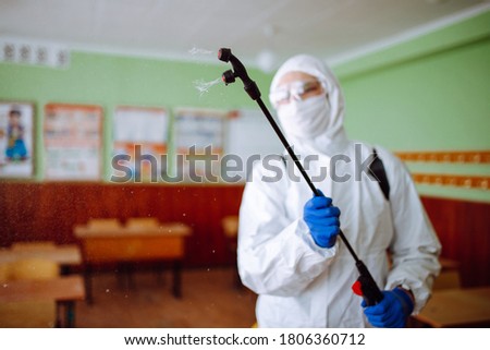Close up of a spray with sanitizing liquid in the hands of a man wearing a protective suit in the schoold classroom. Disinfectant sterilizes the auditorium before the studing process begins. Covid-19.