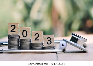 close up stack of coin, 2023 wooden text block and stethoscope on table, saving and manage money for healthcare, life insurance business technology, economic crisis risk and problem concept