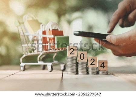 close up stack of coin, 2024 wooden text block, toy shopping cart and small bag on table, hand holding smartphone background, saving and manage money for celebrate season, economic crisis problem