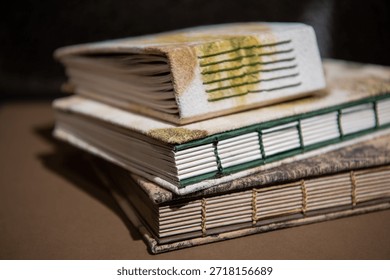 Close up of a stack of handcrafted eco-print notebooks with floral and leaf-patterned covers. Artistic hand-sewn binding and organic textures in a dark studio setting.